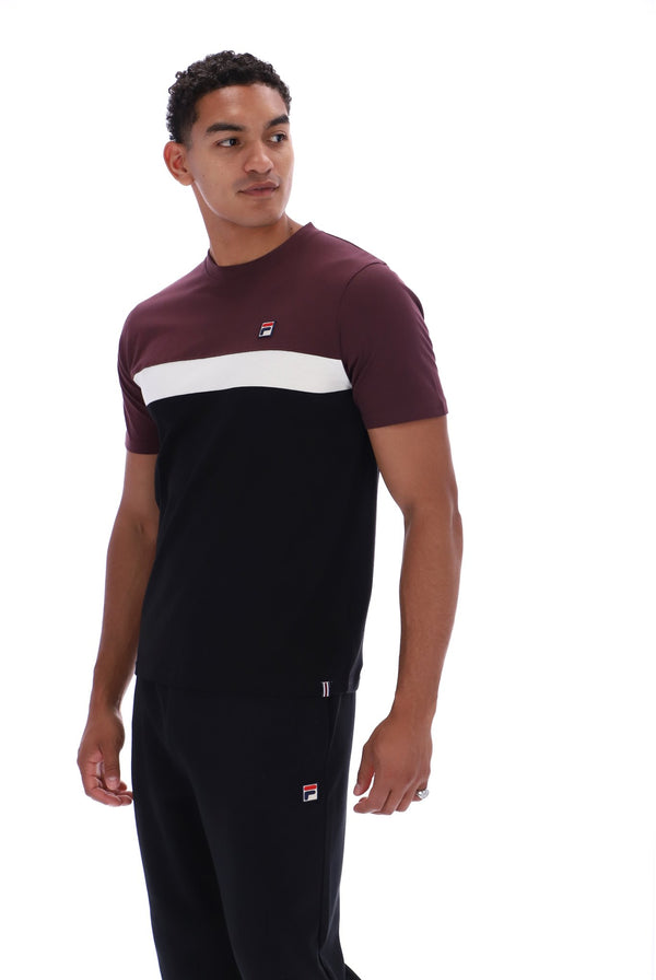 A man wearing a color-blocked short-sleeve t-shirt and matching sweatpants by Fila. The t-shirt features a maroon upper chest and shoulders, a wide white horizontal stripe across the chest, and a solid black lower body.