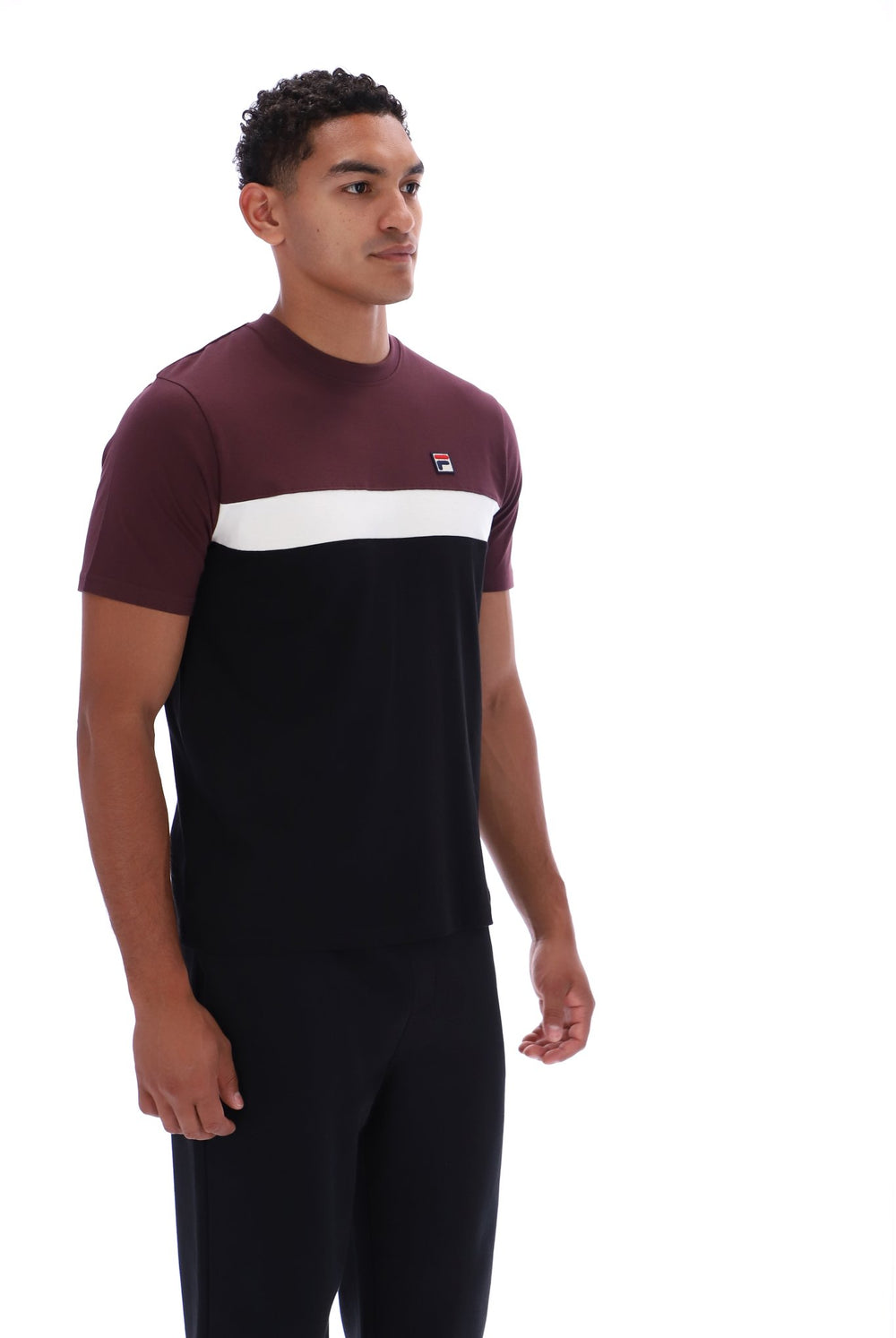 A man wearing a color-blocked short-sleeve t-shirt and matching dark trousers, standing against a plain white studio background. The t-shirt features a maroon upper section, a white horizontal stripe across the chest, and a small red and blue logo on the left chest.