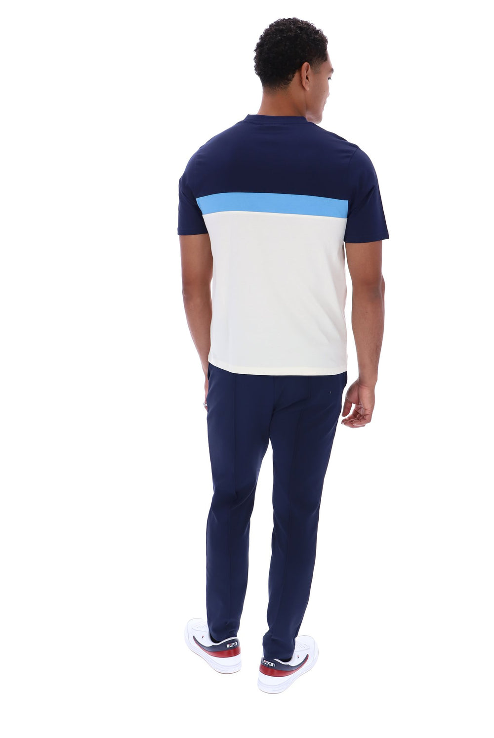 A man viewed from behind wearing a short-sleeve colorblock t-shirt with navy blue, light blue, and white blocks, paired with navy pants and white sneakers.