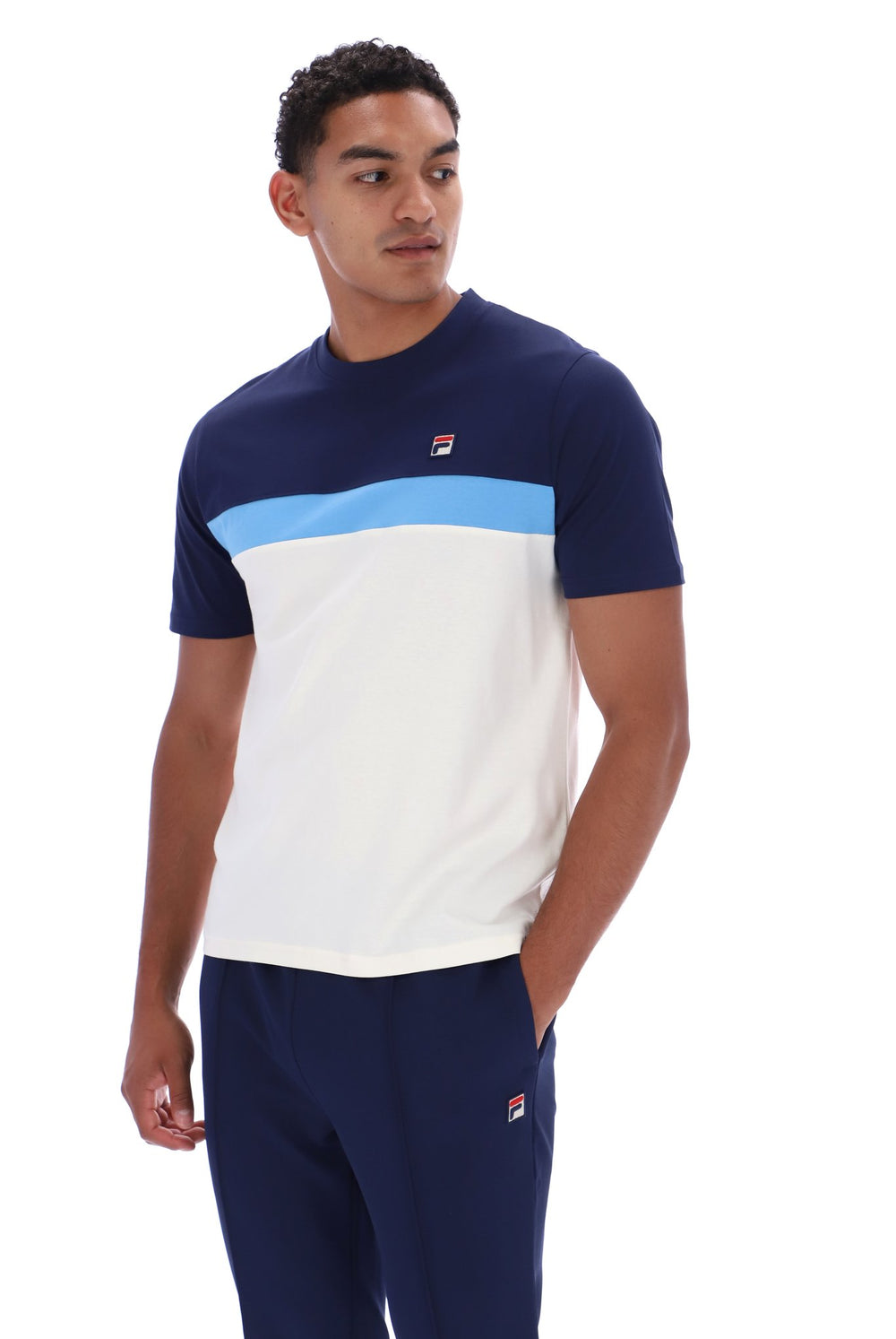 A man wearing a navy blue, white, and light blue color-blocked short-sleeve t-shirt with a small red and white Fila logo on the chest, paired with matching navy pants, against a plain white background.