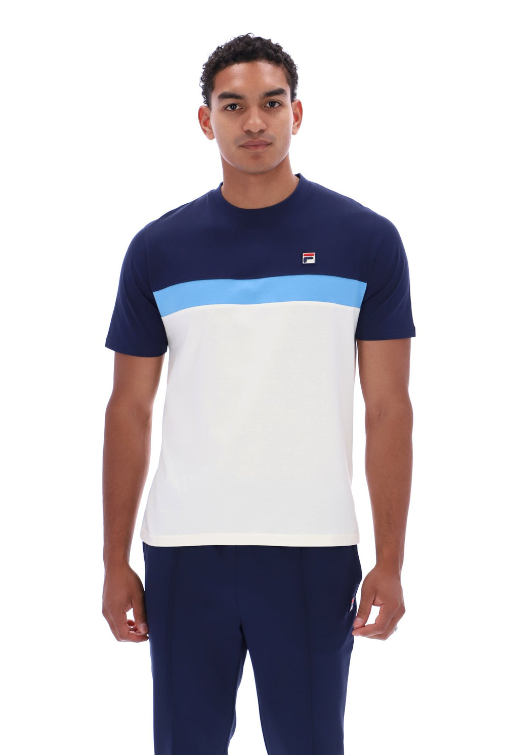 A man wearing a color-blocked short-sleeve t-shirt by FILA in navy blue, light blue, and white, paired with navy pants, against a plain white background.