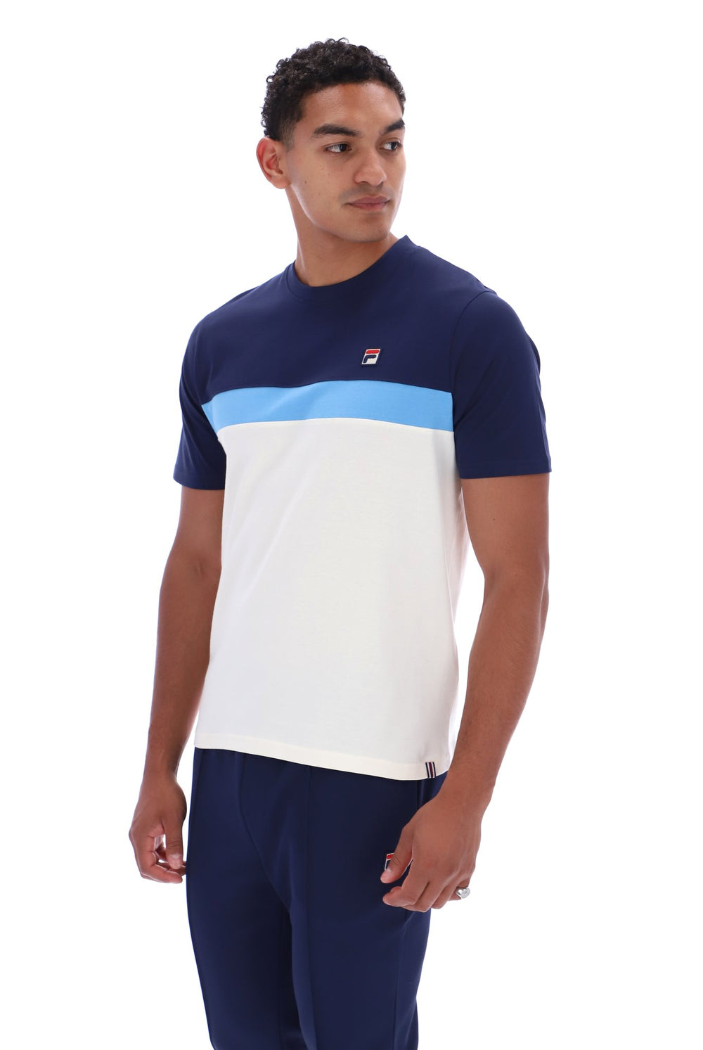 A man wearing a color-blocked short-sleeve t-shirt by FILA in navy blue, white, and light blue, paired with navy pants, against a plain white background.
