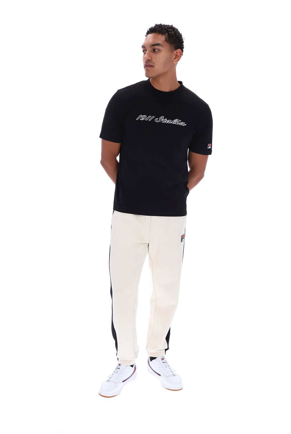 A man modeling a black Champion t-shirt with '1911 Italia' printed on the chest, paired with off-white track pants featuring black side stripes and white sneakers, shot against a solid white background.