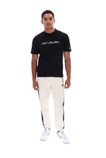 A man modeling a black crewneck t-shirt with white script text on the chest, paired with off-white track pants featuring black side stripes and white sneakers, shot against a solid white background.