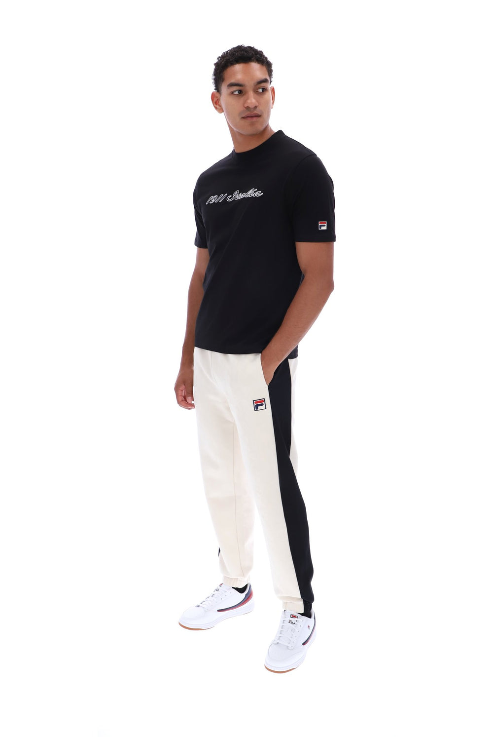 A man modeling a black t-shirt and off-white track pants with black side panels, featuring the Fila logo on the sleeve and thigh.