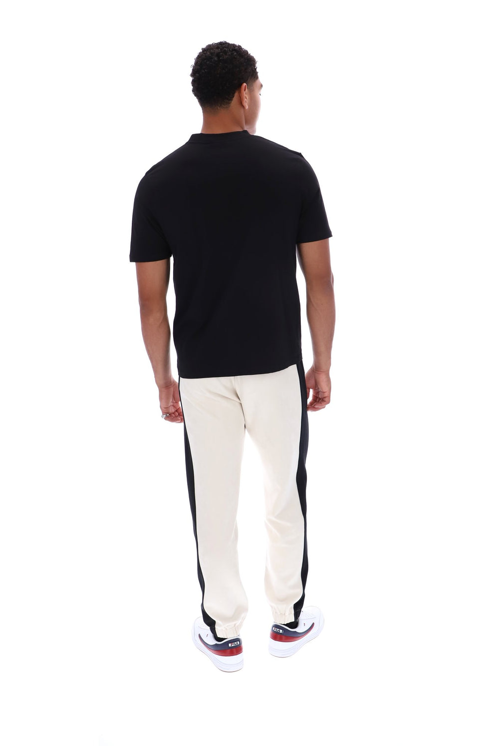 A man viewed from behind wearing a black short-sleeve t-shirt and off-white track pants with black side panels, standing against a plain white background.