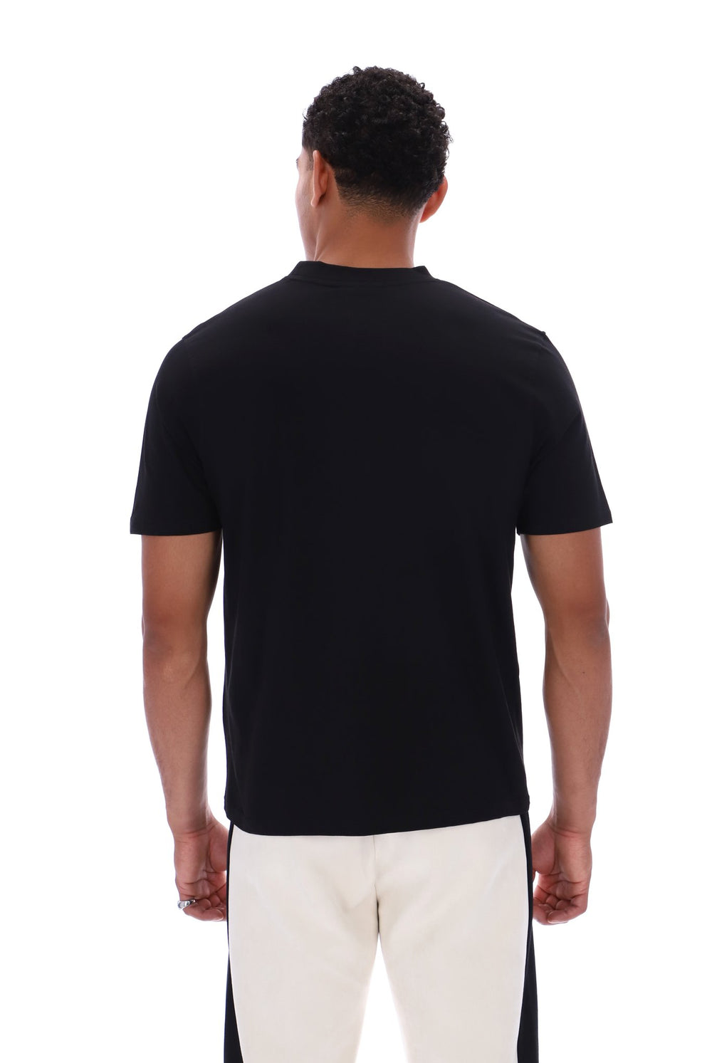 A man viewed from the back wearing a plain black short-sleeve crew-neck t-shirt and off-white pants with black side panels, against a solid white background.