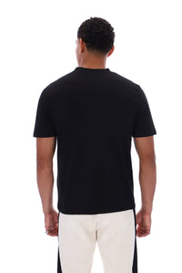 A man viewed from the back wearing a plain black short-sleeve crew-neck t-shirt and off-white pants with black side panels, against a solid white background.
