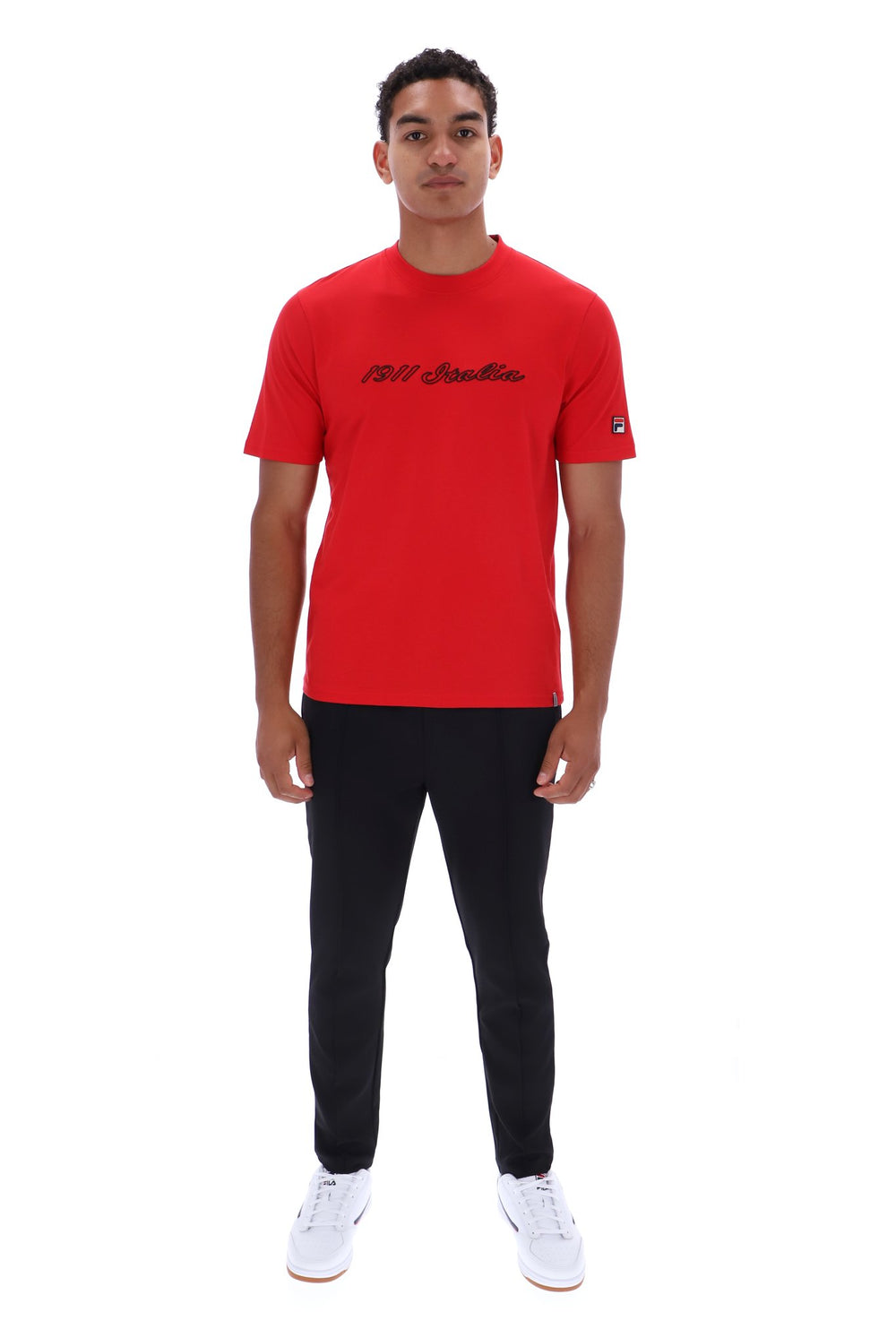 A man modeling a bright red short-sleeve t-shirt with black script text across the chest, paired with black pants and white sneakers, against a plain white background.