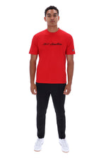 A man modeling a bright red short-sleeve t-shirt with black script text across the chest, paired with black pants and white sneakers, against a plain white background.