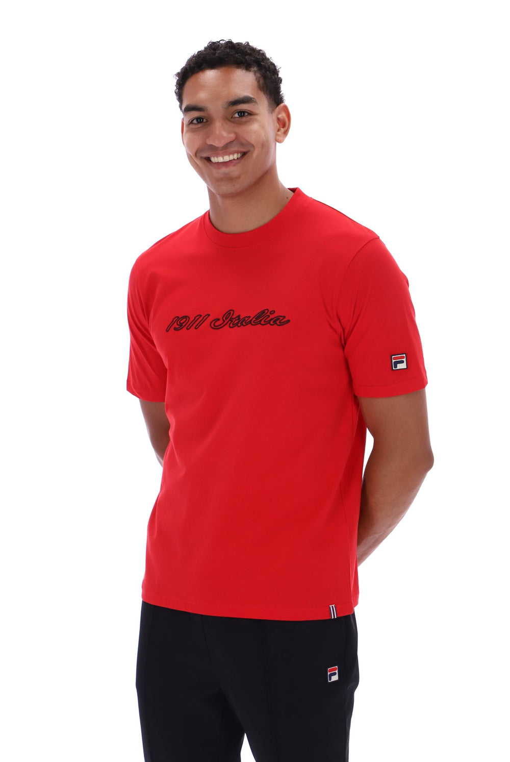 A smiling man wearing a bright red short-sleeve t-shirt with '1911 Italia' printed in black script across the chest, paired with black pants. The t-shirt features a small Fila logo on the left sleeve.