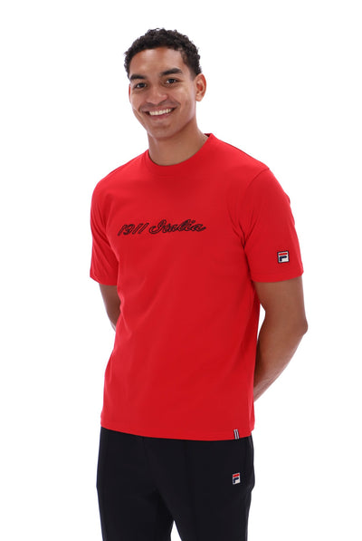 A smiling man wearing a bright red short-sleeve t-shirt with '1911 Italia' printed in black script across the chest, paired with black pants. The t-shirt features a small Fila logo on the left sleeve.
