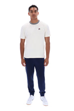 A man modeling a white short-sleeve crewneck t-shirt with a gray ribbed collar and a small red and blue Fila logo on the chest, paired with navy joggers and white sneakers, against a plain white background.