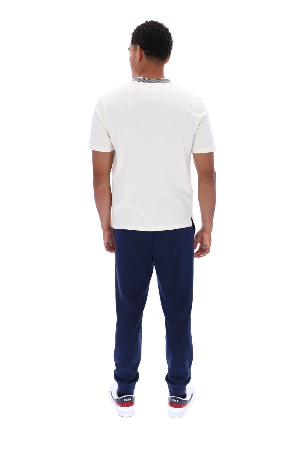 A man viewed from behind wearing a white short-sleeve crewneck t-shirt with a gray collar trim, navy blue pants, and white sneakers with red accents, shot against a solid white background.
