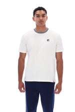 A man wearing a white short-sleeve crewneck t-shirt with a gray ribbed collar and a small Fila logo on the chest, paired with navy pants, against a plain white background.