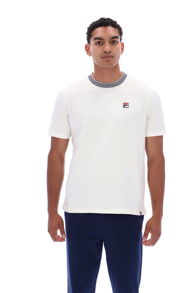 A man wearing a white short-sleeve crewneck t-shirt with a gray ribbed collar and a small Fila logo on the chest, paired with navy pants, against a plain white background.