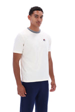 A man wearing a white short-sleeve crewneck t-shirt with a gray ribbed collar and a small red, white, and blue Fila logo on the chest, paired with navy blue pants, against a plain white background.