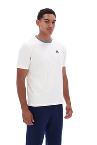 A man wearing a white short-sleeve crewneck t-shirt with a gray ribbed collar and a small red, white, and blue Fila logo on the chest, paired with navy blue pants, against a plain white background.
