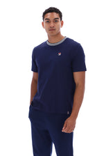 A man wearing a navy blue short-sleeve t-shirt with a gray ribbed crew neck and a small red, white, and blue Fila logo on the left chest, paired with matching navy pants, against a plain white background.