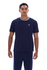A man wearing a navy blue short-sleeve t-shirt with a gray crew neck and a small Fila logo on the chest, paired with matching navy pants, against a plain white background.