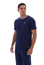 A man wearing a navy blue short-sleeve t-shirt with a gray ribbed crew neck and a small red, white, and blue Fila logo on the left chest, paired with matching navy pants, against a plain white background.