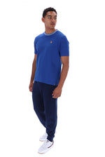 A man modeling a blue short-sleeve crewneck t-shirt with a small red and white logo on the left chest, paired with navy sweatpants and white sneakers, against a plain white background.