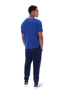 A man shown from the back wearing a royal blue short-sleeve t-shirt, navy blue jogger pants, and white sneakers with red accents, set against a plain white background.