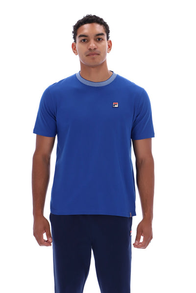 A man wearing a blue short-sleeve crewneck t-shirt with a gray collar trim and a small red, white, and blue Fila logo on the left chest, paired with navy pants, against a plain white background.