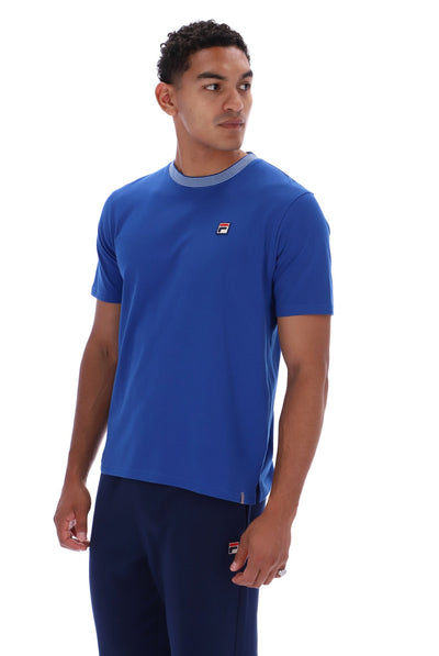 A man wearing a blue short-sleeve crewneck t-shirt with a small Fila logo on the chest, paired with navy pants, photographed against a plain white background.