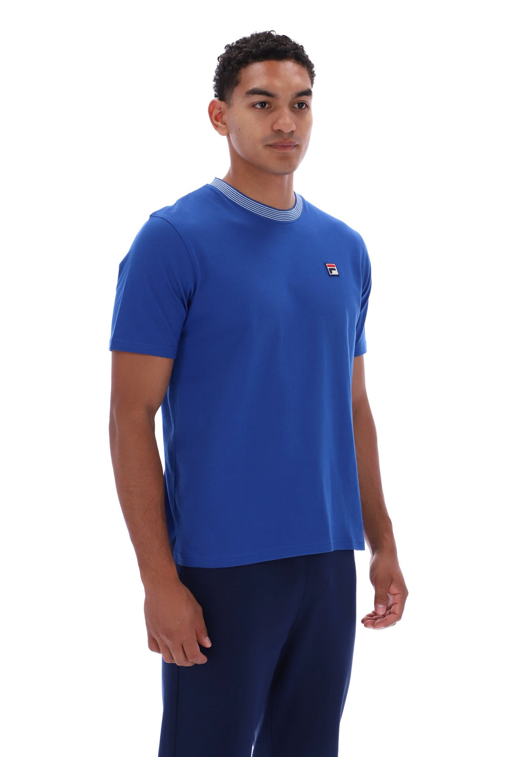 A man wearing a royal blue short-sleeve crewneck t-shirt with a light gray ribbed collar and a small red, white, and blue logo on the left chest, paired with navy blue pants, against a plain white background.