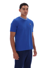 A man wearing a royal blue short-sleeve crewneck t-shirt with a light gray ribbed collar and a small red, white, and blue logo on the left chest, paired with navy blue pants, against a plain white background.