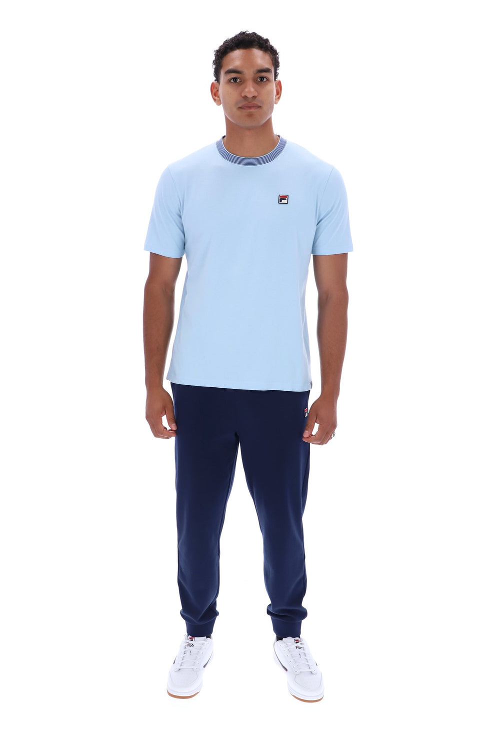 A man modeling a light blue short-sleeve crewneck t-shirt with a small red and white Fila logo on the left chest, paired with navy joggers and white sneakers, against a plain white background.
