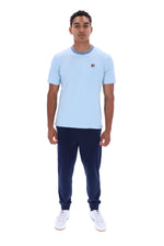 A man modeling a light blue short-sleeve crewneck t-shirt with a small red and white Fila logo on the left chest, paired with navy joggers and white sneakers, against a plain white background.