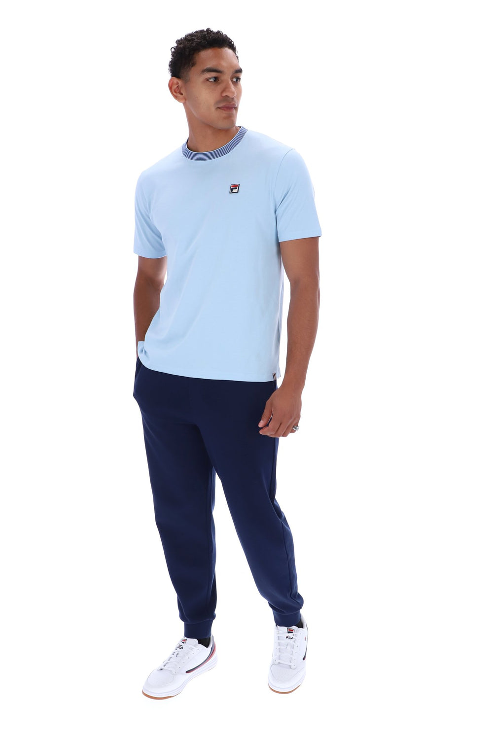 A man modeling a light blue short-sleeve t-shirt with a dark blue crew neck collar and a small red and white Fila logo on the chest, paired with navy joggers and white sneakers, against a plain white background.