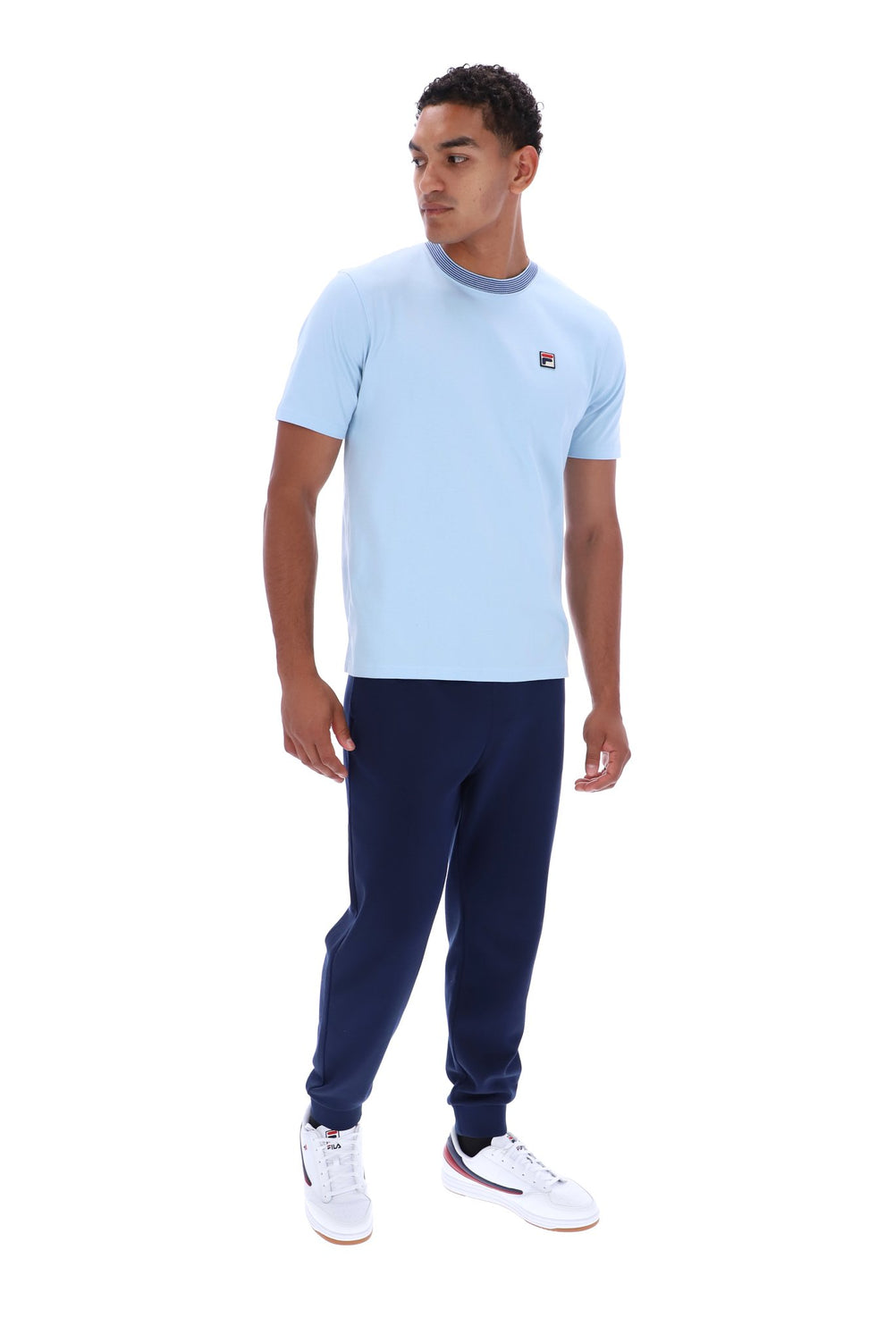 A man modeling a light blue short-sleeve crewneck t-shirt with a small red and white logo on the left chest, paired with navy joggers and white sneakers, against a plain white background.