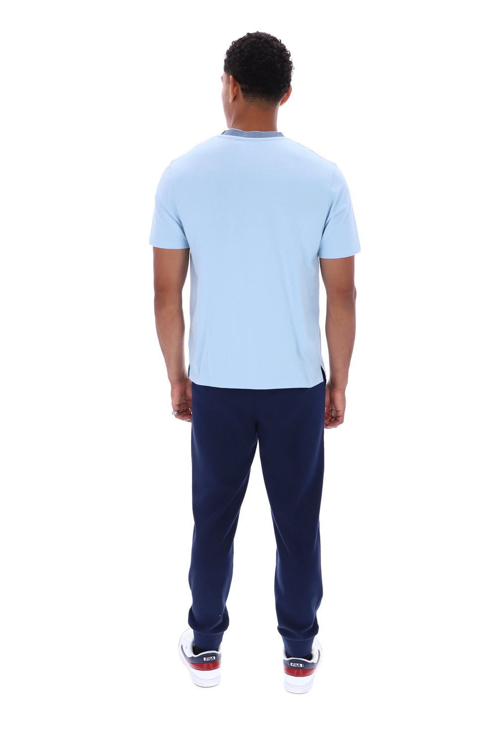 A man viewed from behind wearing a light blue short-sleeve t-shirt and navy blue sweatpants, standing against a solid white background.