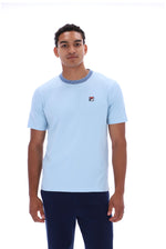 A man wearing a light blue short-sleeve crewneck t-shirt with a contrasting darker blue collar and a small red, white, and blue Fila logo on the left chest, paired with dark navy pants, against a plain white background.