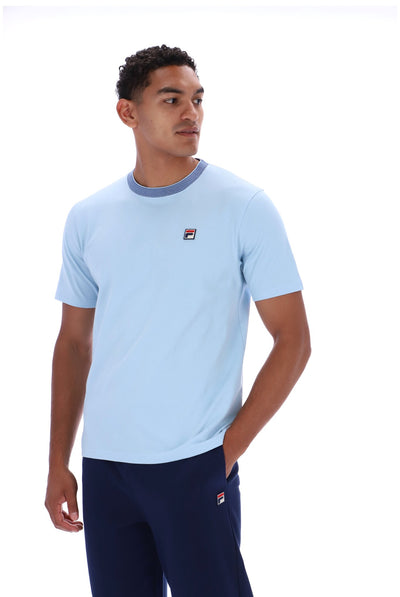 A man wearing a light blue short-sleeve crewneck t-shirt with a small red, white, and blue Fila logo on the left chest, paired with navy blue pants, against a plain white background.