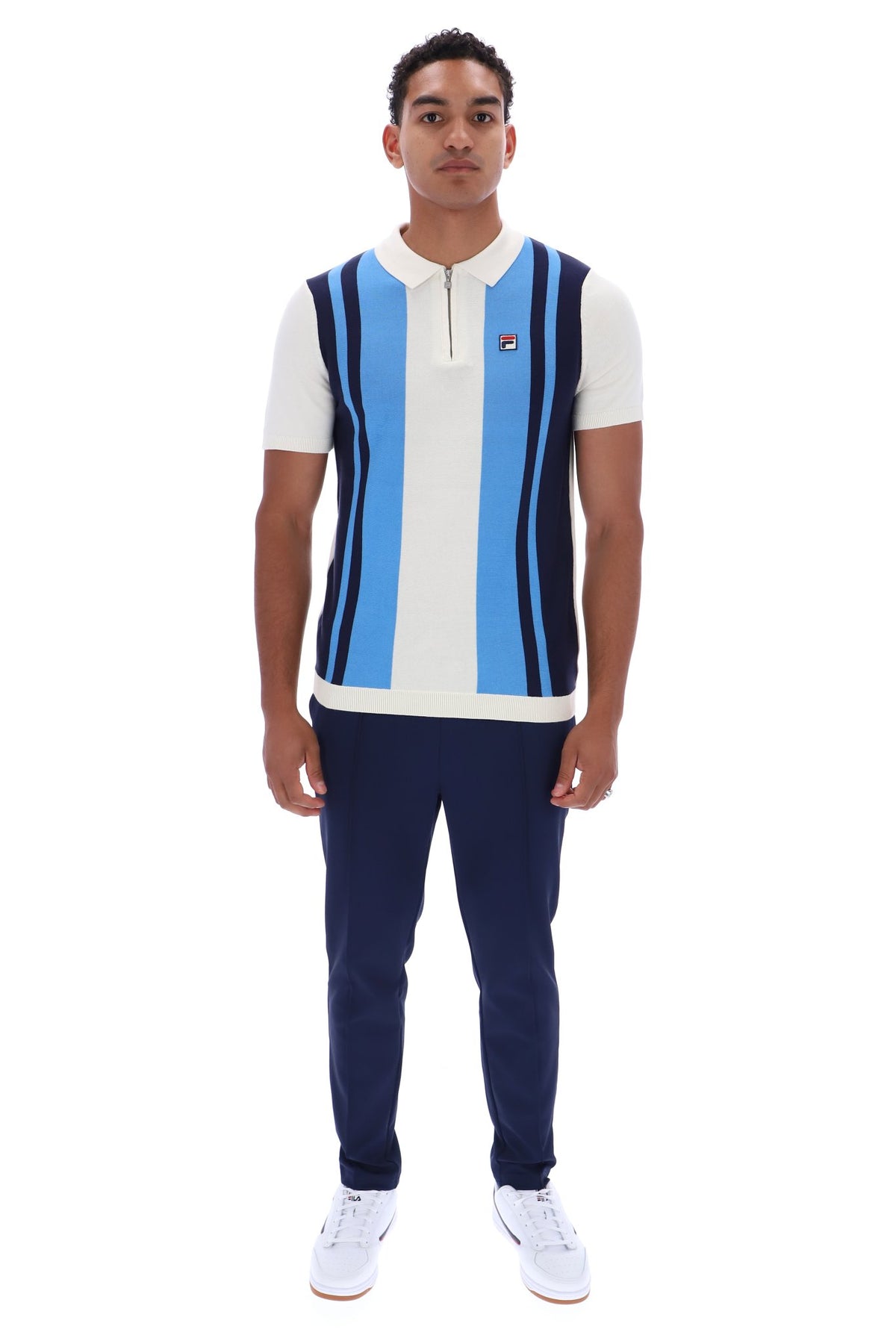 A man modeling a FILA short-sleeve polo knit vest with vertical light blue, white, and navy stripes, paired with navy pants and white sneakers, on a solid white background.