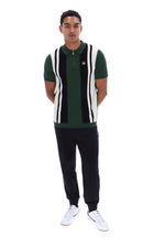 A man modeling a green, black, and white striped knit polo vest over a green polo shirt, paired with black sweatpants and white sneakers, against a plain white background.