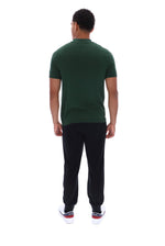 A man viewed from behind wearing a dark green short-sleeve polo shirt, black pants, and white sneakers with red accents, set against a plain white background.
