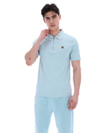 A man wearing a light blue zip-up polo shirt with a FILA gold logo on the chest, on a white background