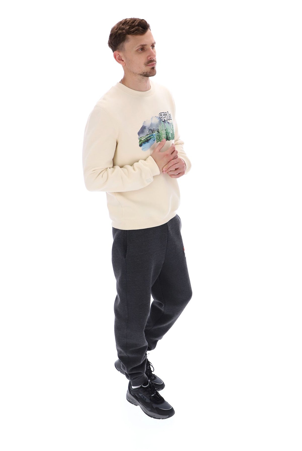 A male model wearing a beige FILA Len Graphic Crew Sweatshirt with a mountain range and brand logo on the front.