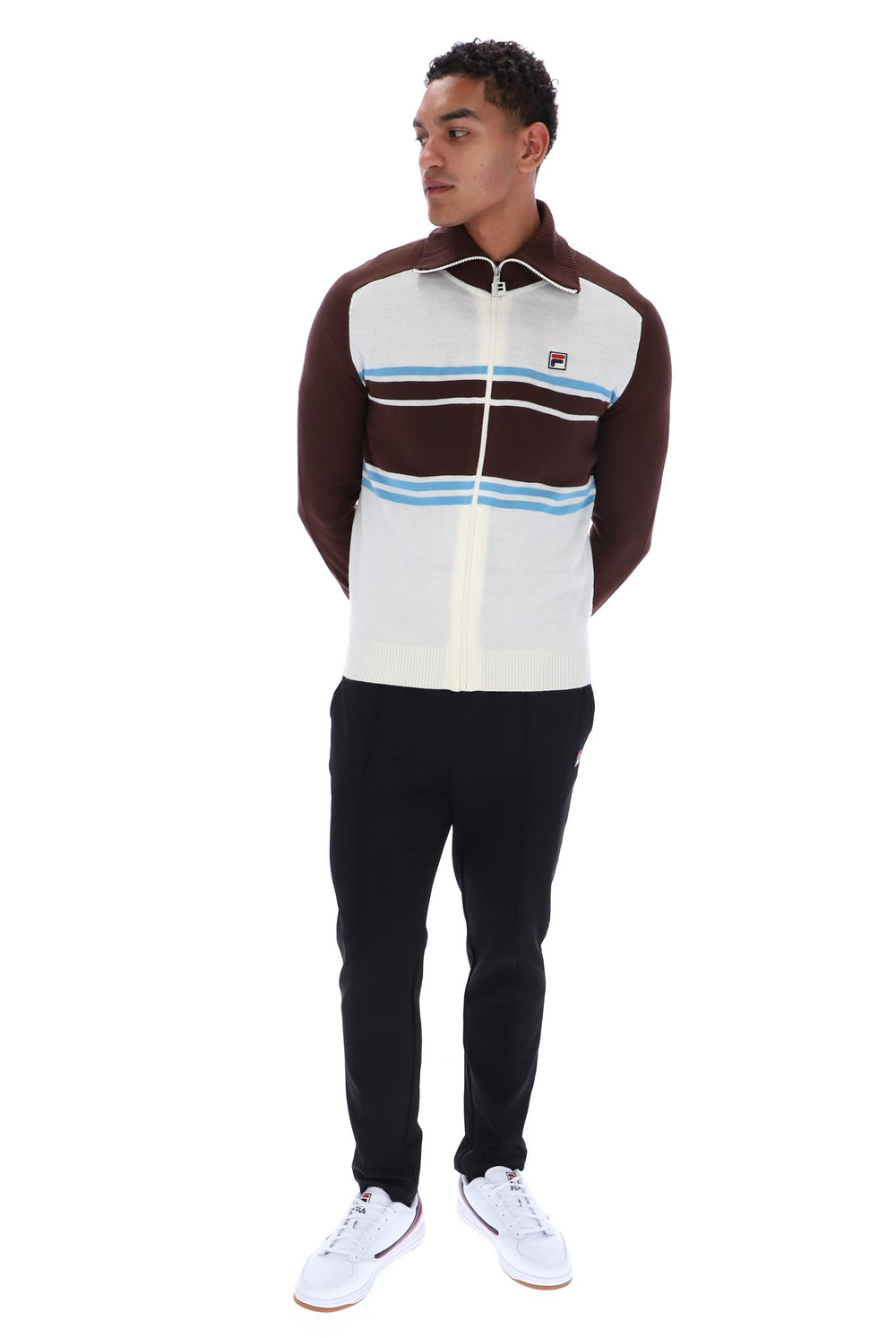 A man modeling a Fila zip-up track jacket with a cream body, brown shoulders and collar, and light blue and white horizontal stripes across the chest, paired with black pants and white sneakers against a white background.