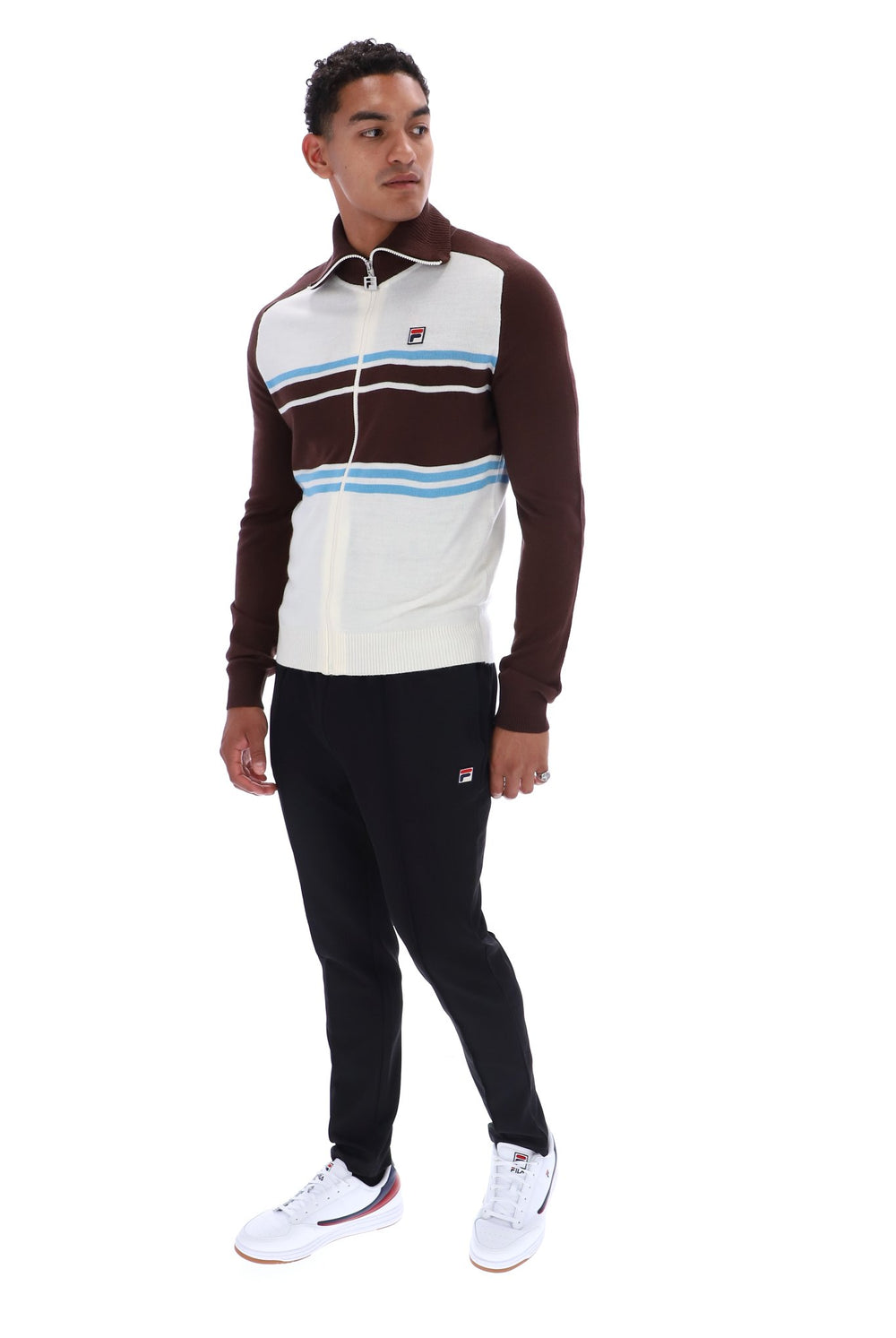 A man modeling a Fila tracksuit featuring a brown and white zip-up jacket with light blue stripes, paired with black joggers and white sneakers, on a solid white background.