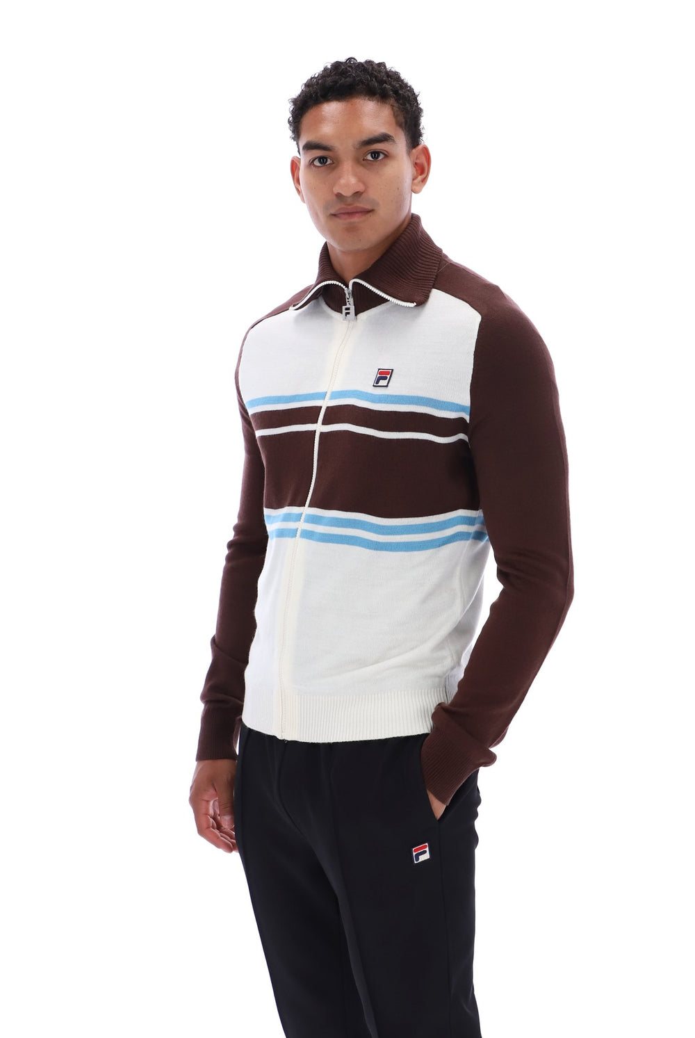 A man wearing a brown and white Fila quarter-zip track jacket with light blue stripes, paired with black sweatpants, against a plain white background.