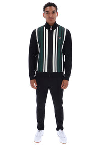 A man modeling a black track jacket with vertical green, white, and black stripes on the front, paired with black sweatpants and white sneakers, against a plain white background.