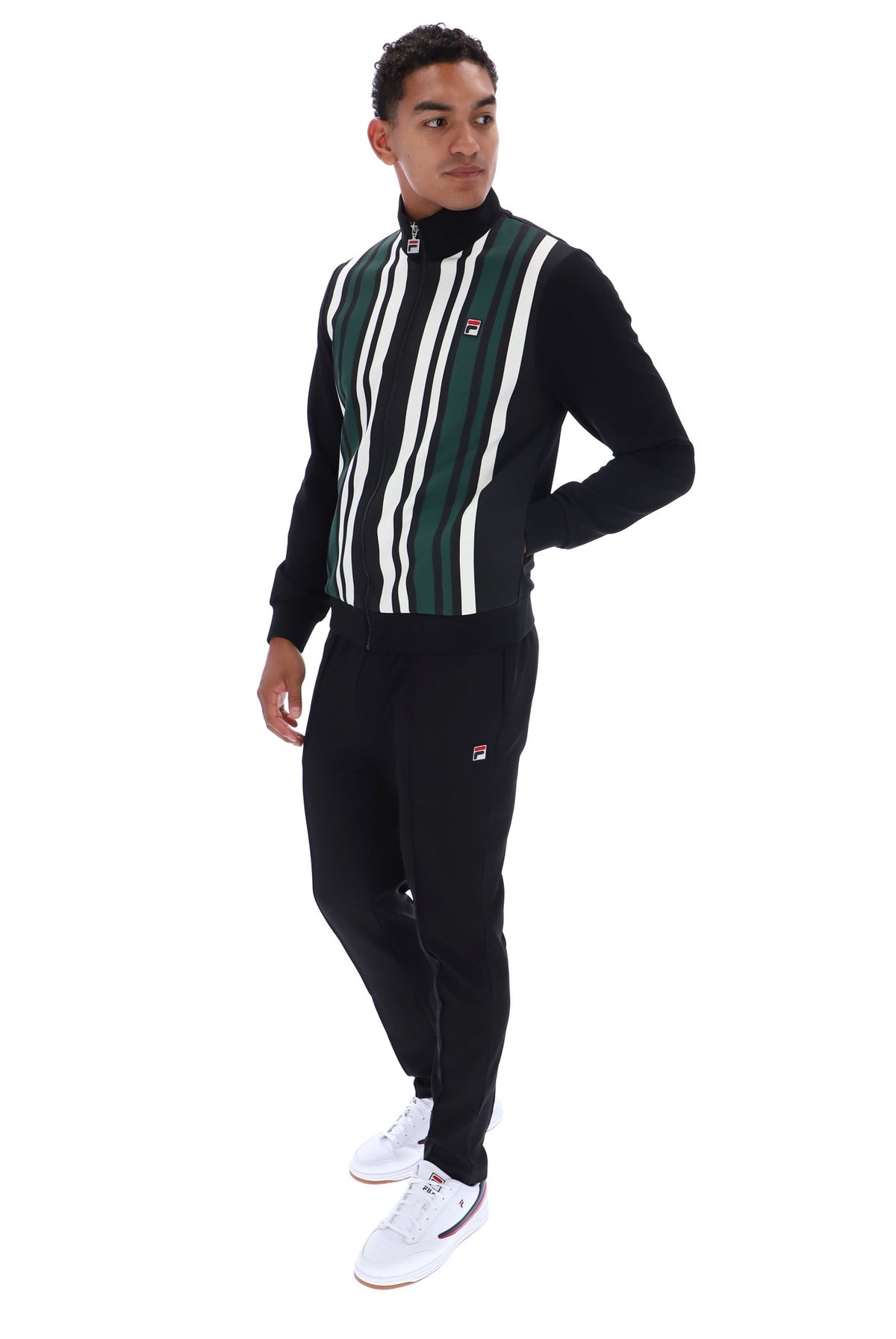 A man modeling a black knit tracksuit with green and white vertical stripes on the chest, featuring a mock neck, zippered collar, and matching black joggers with a small red and white logo.