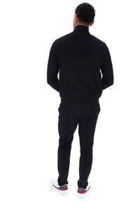 A man viewed from behind wearing a black long-sleeve mock-neck top and matching black pants, paired with white sneakers that have red and black accents, against a plain white background.