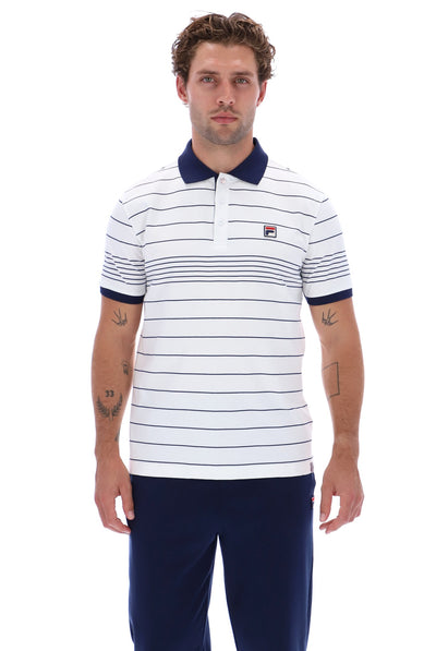 A man wearing a white short-sleeve polo shirt with thin navy horizontal stripes, a navy collar, and a small red and white Fila logo on the chest, paired with navy pants, against a plain white background.